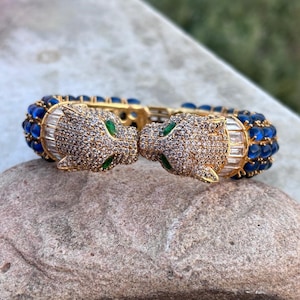 May include: A gold-tone hinged bracelet featuring two panther heads facing each other. The panther heads are encrusted with clear pavé stones and green stone eyes. The band is decorated with rows of blue cabochon stones and clear baguette crystals.