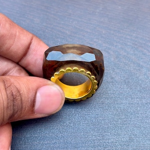 以下が含まれることがあります： ファセット加工された、スモーキーブラウンの宝石リング、ゴールドバンド付き。 リングは幾何学的な形状で、ゴールドバンドにはスカラップエッジがあります。 宝石はゴールドバンドで固定されています。