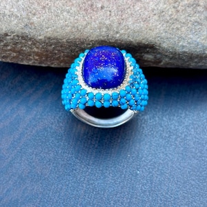 May include: A silver ring featuring a large, oval, deep blue gemstone at the center, surrounded by a halo of small, turquoise-colored beads. The ring's band is a polished silver.