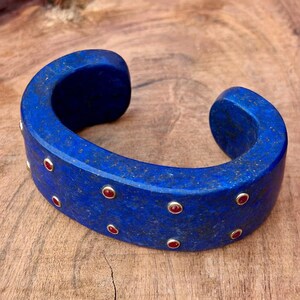 May include: A striking blue cuff bracelet with a curved shape. The bracelet features small, round red gemstones set in gold-coloured bezels. The bracelet is presented on a wooden surface.