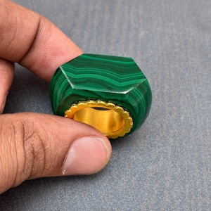 以下が含まれることがあります： 独特の縞模様を持つ、大きなファセットカットの濃い緑色のマラカイト石をあしらったリング。石は光沢のあるゴールドカラーの金属バンドにはめ込まれており、リングの内周には装飾的なスカラップ状の縁取りが施されています。