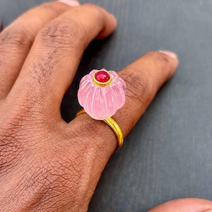 May include: A gold ring featuring a carved, translucent, light pink, pumpkin-shaped gemstone. The center of the pink stone is accented with a small, round, vibrant red gemstone set in a gold bezel. The ring band is a simple, polished gold metal.