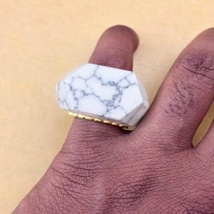 以下が含まれることがあります： グレーの脈状模様が入った大きなファセットカットの白い石をあしらった指輪。石はゴールドトーンの金属バンドに取り付けられており、セッティングの下側にはスカラップ状またはテクスチャー加工された縁が見えます。