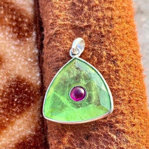 May include: A triangular pendant with a silver bail. The pendant features a large, faceted, light green gemstone with a smaller, round, deep pink gemstone set in the center. The pendant is displayed on a brown, textured surface.