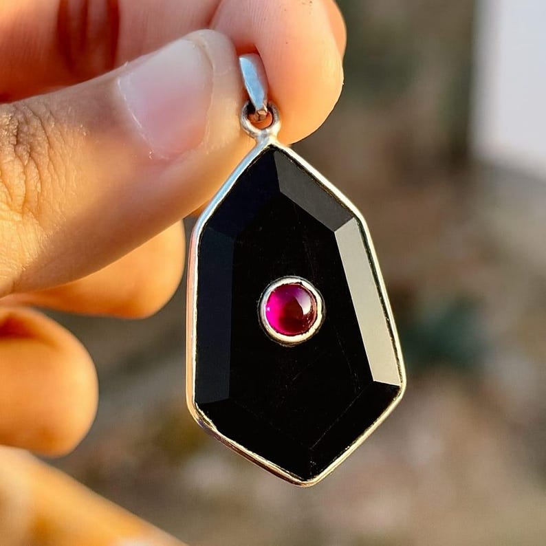 May include: A black gemstone pendant with a geometric shape and a small, round, red gemstone in the center. The pendant has a silver-colored metal border and a silver loop at the top for a chain.