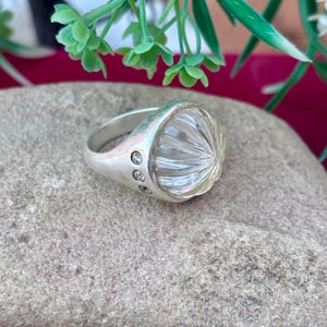 May include: A silver ring featuring a large, clear, faceted gemstone. The band has three small, clear stones set into it. The ring is shown on a grey stone surface, with green foliage in the background.