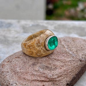 Pode incluir: Um anel com uma banda feita de coral fossilizado bege texturizado com um padrão de estrela. A banda apresenta uma única pedra preciosa verde vibrante, lapidada em almofada facetada, fixada numa moldura de metal prateado liso e polido.