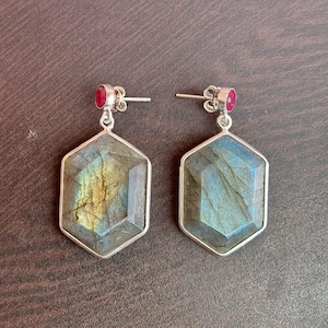 May include: A pair of silver earrings featuring hexagonal labradorite stones with iridescent blue and green hues. Each earring has a small, round, red gemstone at the top.