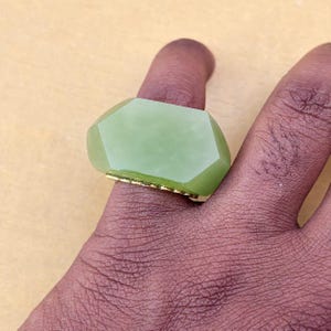 以下が含まれることがあります： 薄いゴールドトーンの金属バンドにセットされた、大きなファセットカットのライトグリーンの石が付いたリング。石は滑らかで磨かれた表面を持ち、端に沿って複数の平らで角度のあるファセットがあり、指の上に幾何学的な形状を作り出しています。