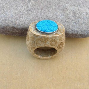 以下が含まれることがあります： ベージュ色のリング。花柄模様と、銀色のベゼルにセットされたターコイズブルーの花型の石が特徴です。リングは明るい色の素材で作られており、指を入れるための大きな開口部がある丸みを帯びた形状をしています。