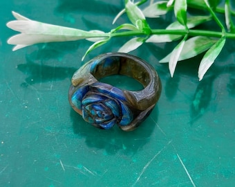 Può includere: Un anello di pietra preziosa scolpita con un motivo a rosa blu. L'anello ha una tonalità scura e terrosa con una finitura lucida. La rosa è finemente dettagliata, con sfumature di blu e verde. L'anello è posto su uno sfondo verde con fiori bianchi.