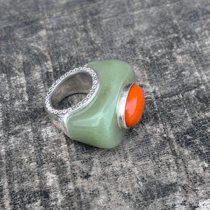 May include: A ring featuring a smooth, light green stone body with a polished silver band edge decorated with small circular indentations. A vibrant, round orange stone is set into the side of the green stone, held in place by a silver bezel setting.