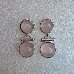Pode incluir: Um par de brincos prateados com um design circular e uma pedra transl&uacute;cida rosa claro. Cada brinco tem duas pedras circulares conectadas por uma pequena barra horizontal. Os brincos s&atilde;o exibidos em uma superf&iacute;cie cinza claro texturizada.