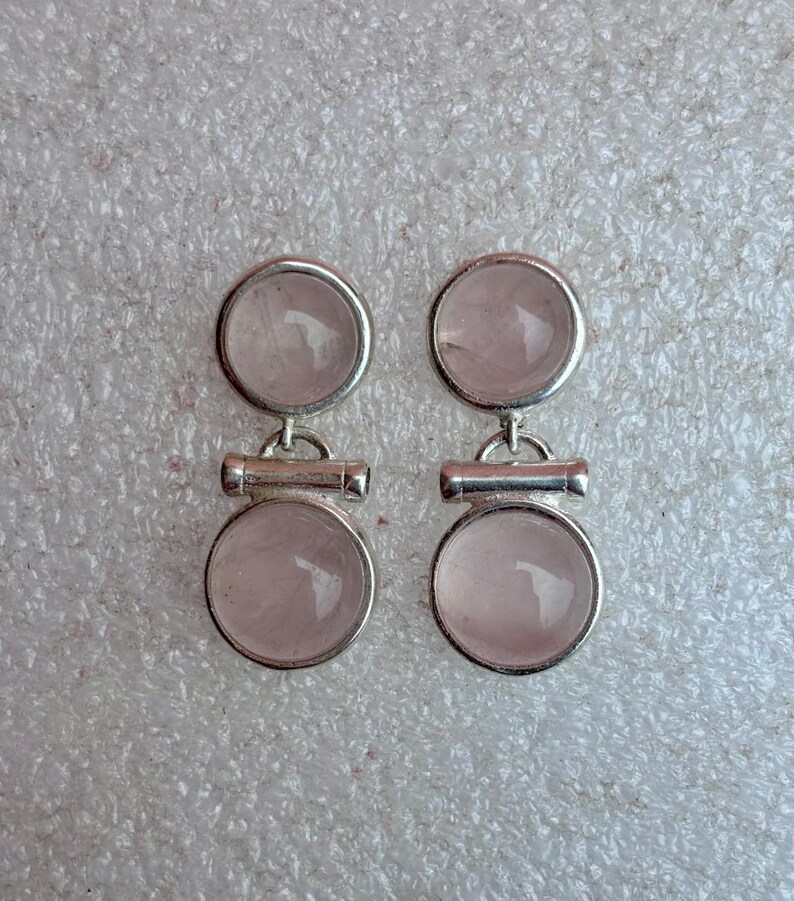 Pode incluir: Um par de brincos prateados com um design circular e uma pedra transl&uacute;cida rosa claro. Cada brinco tem duas pedras circulares conectadas por uma pequena barra horizontal. Os brincos s&atilde;o exibidos em uma superf&iacute;cie cinza claro texturizada.