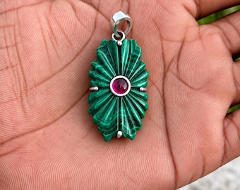 May include: A pendant featuring a carved green malachite stone with a radial, fan-like pattern and a central circular magenta gemstone. The piece is set in a silver-toned metal frame with a top bail for attachment to a chain or cord.