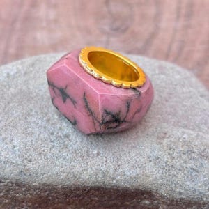以下が含まれることがあります： ゴールドバンドのピンク色の宝石リング。リングは、黒い静脈のあるファセット加工されたピンク色の宝石にセットされています。ゴールドバンドはスカラップエッジです。リングは灰色の表面に表示されます。