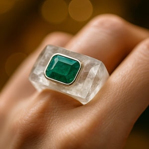 May include: A rectangular ring with a large, emerald-cut green gemstone set in a gold bezel. The gemstone is set in a clear, rectangular base. The ring is on a person's finger.