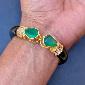 Könnte beinhalten: Ein schwarzes und goldenes Armreif-Armband mit zwei tropfenförmigen grünen Edelsteinen. Das Armband hat goldfarbene Akzente und klare, rechteckig geschliffene Steine. Das Armband befindet sich an einem Handgelenk.
