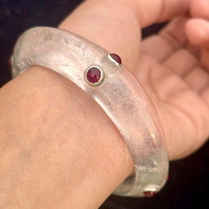 May include: A clear, translucent bangle bracelet with three small, round, red gemstones. The bracelet is worn on a wrist, highlighting its smooth, curved design. The gemstones are evenly spaced around the bracelet.