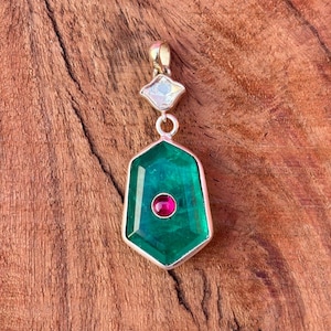 May include: A silver pendant with a large, emerald-green gemstone in a hexagonal shape. A small, round, red gemstone is set in the center. The pendant is suspended from a smaller, diamond-shaped, iridescent stone.