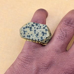 以下が含まれることがあります： ダルメシアンのような黒い斑点模様が入った、大きなベージュ色のファセットカットストーンがあしらわれた指輪。ストーンはゴールドトーンの金属製バンドの上にセットされており、指輪のファセット加工された上部の下からそのバンドが見えるデザインです。