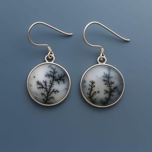 May include: A pair of round silver earrings. Each earring features a white and gray stone with a black, tree-like pattern. The earrings have a simple hook design for pierced ears. The background is a solid, muted blue.