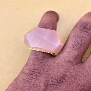 以下が含まれることがあります： ゴールドトーンの金属バンドにセットされた、大きなファセットカットの淡いピンク色の宝石が付いた指輪。石は滑らかで磨かれた表面を持つ幾何学的な多面カットが施されています。石の下には金属製のゴールドのバンドが見えています。