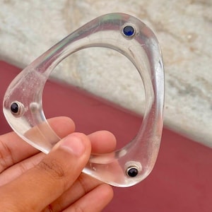 Könnte beinhalten: Transparentes, dreieckiges Armband mit drei kleinen, runden, dunklen Edelsteinen. Das Armband wird in einer Hand gehalten, und der Hintergrund ist eine Mischung aus Rosa und Weiß.