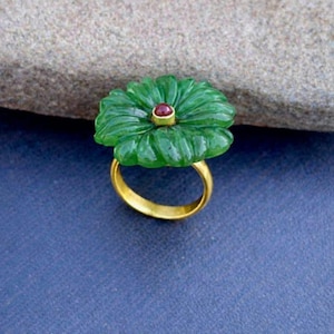 May include: A gold ring featuring a carved green gemstone in the shape of a flower. The flower has a small red gemstone in the center. The ring is set against a blue background.