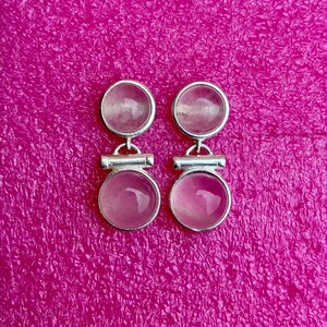 Pode incluir: Um par de brincos prateados com design de pedras rosas. Cada brinco apresenta uma pedra pequena e redonda no topo e uma pedra rosa circular maior em baixo, conectadas por uma barra horizontal. Fundo magenta texturizado.