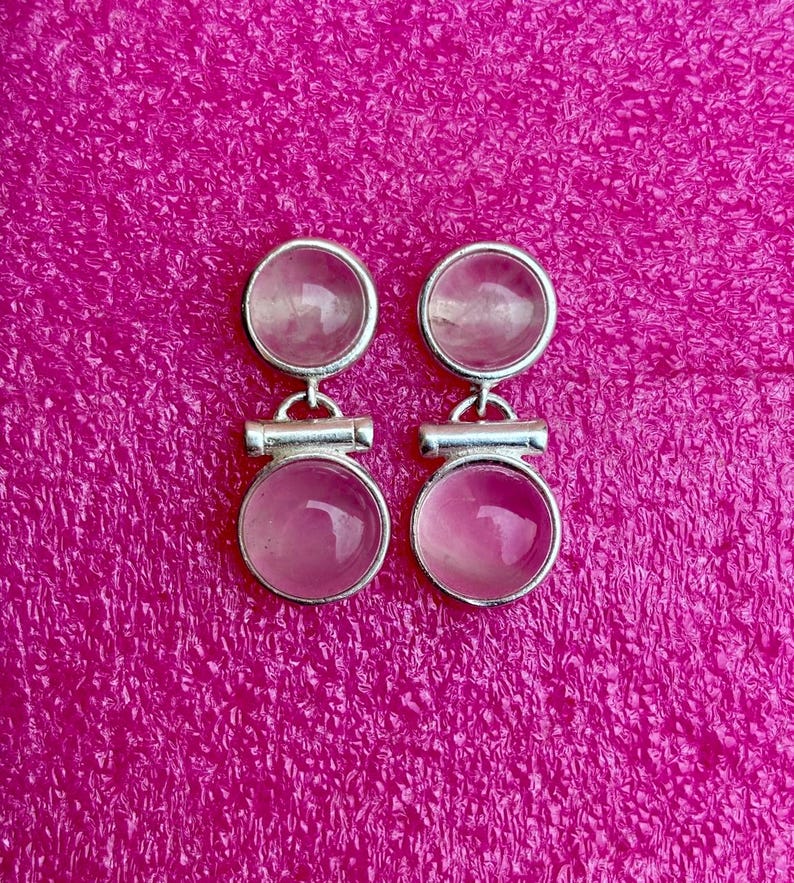 Pode incluir: Um par de brincos prateados com design de pedras rosas. Cada brinco apresenta uma pedra pequena e redonda no topo e uma pedra rosa circular maior em baixo, conectadas por uma barra horizontal. Fundo magenta texturizado.