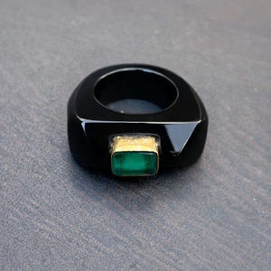 以下が含まれることがあります： 長方形のエメラルドグリーンの宝石が金色のベゼルにセットされた黒い指輪。指輪は、研磨仕上げの厚い正方形のバンドが付いています。宝石の長さは約0.6cmです。