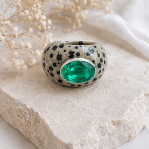 Può includere: Un anello largo caratterizzato da un motivo maculato beige e nero sulla fascia, centrato da una grande pietra preziosa verde brillante a taglio ovale, incastonata in una lunetta liscia color argento. L'anello ha un profilo spesso e arrotondato.