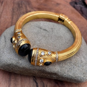 Peut inclure: Un bracelet jonc articulé doré orné de deux pierres cabochons noires proéminentes aux extrémités ouvertes. La surface métallique présente des détails nervurés et est agrémentée de petits cristaux clairs sertis pavé autour des pierres noires.