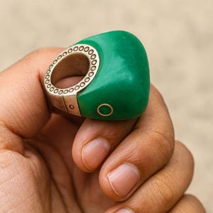 May include: A unique ring featuring a vibrant green, rounded top and a silver band with a series of small circular cutouts. The ring has a modern design and is worn on a hand.