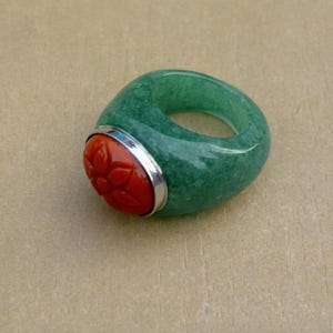 以下が含まれることがあります： 彫刻された赤い花の模様が特徴の、分厚い緑色の指輪。指輪は赤い石の周りに銀色のバンドが付いています。指輪は滑らかで光沢のある素材で作られています。