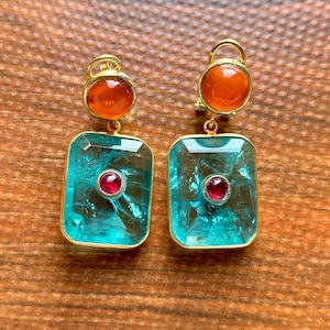 Peut inclure: Une paire de boucles d'oreilles dorées. Chaque boucle d'oreille présente une pierre ronde, rouge orangé en haut et une pierre rectangulaire, bleu-vert avec une petite pierre rouge ronde au centre. Les boucles d'oreilles sont posées sur une surface en bois.