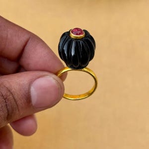 以下が含まれることがあります： 垂直の溝が彫られた大きな球状の黒い石をあしらったゴールドの指輪。黒い石の頂部には、滑らかで丸いピンクがかった赤色の宝石がゴールドの枠にはめ込まれており、色と質感の鮮やかなコントラストを生み出しています。