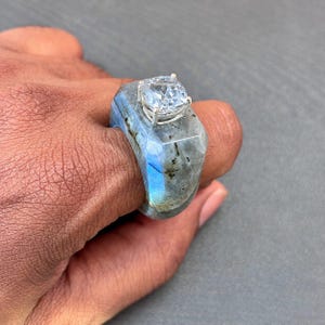以下が含まれることがあります： 青い虹色の光沢と暗いインクルージョンを持つ、磨かれたファセットカットのラブラドライトバンドの指輪。厚みのある石のバンドの上部には、4本の爪を持つ銀色の金属製の台座に、透明なスクエアカットの宝石がセットされています。