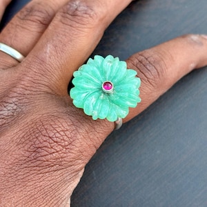 May include: A silver ring featuring a carved, light green flower design with a small, round, red gemstone at the center. The ring is worn on a finger, showcasing the intricate details of the floral motif.
