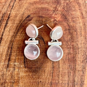 Pode incluir: Um par de brincos de quartzo rosa. Cada brinco apresenta uma pedra redonda rosa claro emoldurada em uma arma&ccedil;&atilde;o de metal prateado. Os brincos t&ecirc;m um design de tarraxa e s&atilde;o exibidos em uma superf&iacute;cie de madeira.