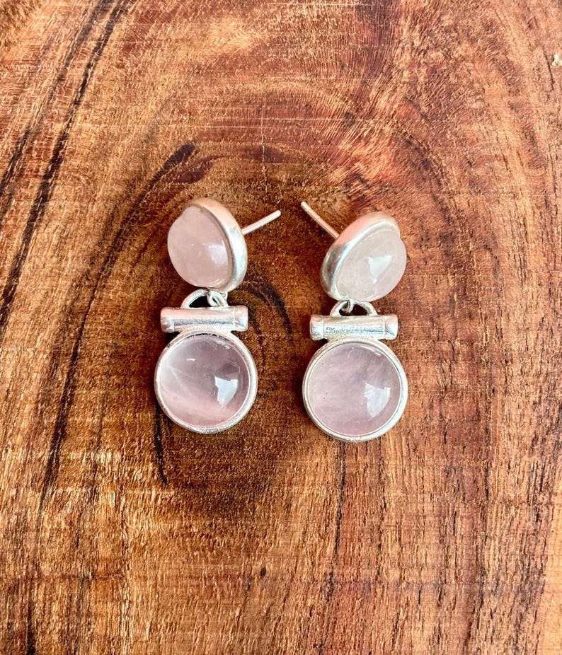 Pode incluir: Um par de brincos de quartzo rosa. Cada brinco apresenta uma pedra redonda rosa claro emoldurada em uma arma&ccedil;&atilde;o de metal prateado. Os brincos t&ecirc;m um design de tarraxa e s&atilde;o exibidos em uma superf&iacute;cie de madeira.