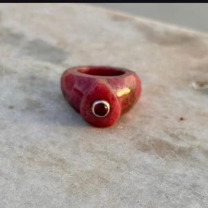 May include: A thick, textured ring crafted from a mottled reddish-pink material. The band features a prominent, raised teardrop-shaped focal point on the front, which is inlaid with a small, circular, dark red stone set in a polished silver bezel.