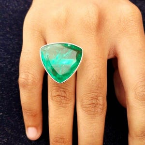 May include: A silver ring featuring a large, triangular, emerald-green gemstone. The gemstone has a faceted surface that catches the light, creating a sparkling effect. The ring's band is a simple, polished silver.