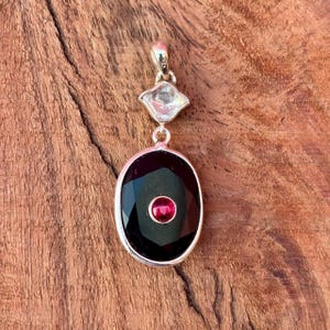 May include: A silver pendant featuring a large, faceted, oval black gemstone, with a small, round, red gemstone at its centre. Above the oval is a clear, diamond-shaped gemstone. The pendant hangs from a silver bail.