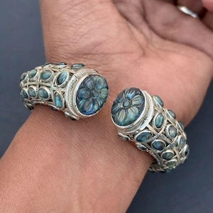May include: Silver cuff bracelet with oval-shaped blue-green stones and carved floral designs. The bracelet features a hinged opening and is designed to be worn on the wrist. The stones are set in a silver metal.