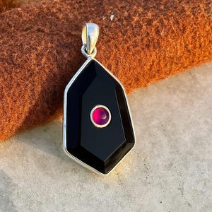 May include: A geometric pendant with a black onyx center and a small, round, pink gemstone. The pendant has a silver-colored bail and a silver-colored border. The pendant is displayed against a brown background.