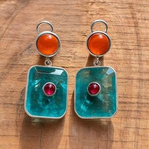 Puede incluir: Un par de pendientes de plata con un diseño único. Cada pendiente presenta una piedra naranja redonda en la parte superior, una piedra turquesa cuadrada en el medio y una pequeña piedra roja en el centro del cuadrado. Sobre una superficie de madera.