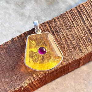 Puede incluir: Un colgante de plata con una gema grande, facetada, de color amarillo-naranja. Una pequeña piedra redonda de color rosa intenso está engastada en el centro. El colgante tiene una fianza de plata en la parte superior para una cadena. La gema está engastada en un bisel de plata.