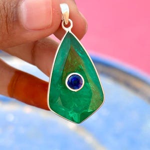 May include: A teardrop-shaped pendant with a large, faceted green gemstone and a small, round, blue gemstone in the center. The pendant is set in a silver metal frame. The gemstone is approximately 1.5 inches long.
